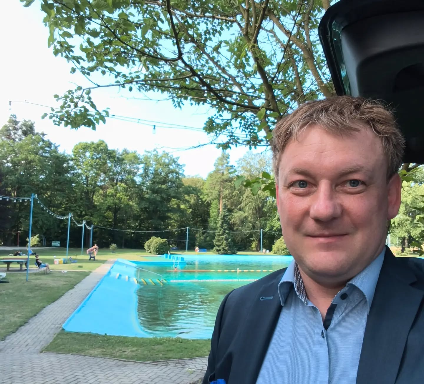 Mann im Vordergrund vor blauem Pool, Bäume und Spielplatz im Hintergrund bei Tageslicht.