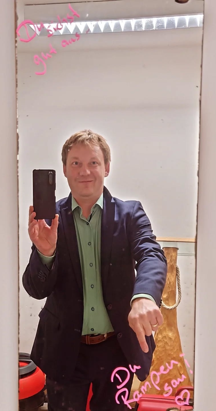 Mann im Anzug hält ein Smartphone vor dem Spiegel. Auf dem Spiegel steht "Du siehst gut aus Du Rampensau".